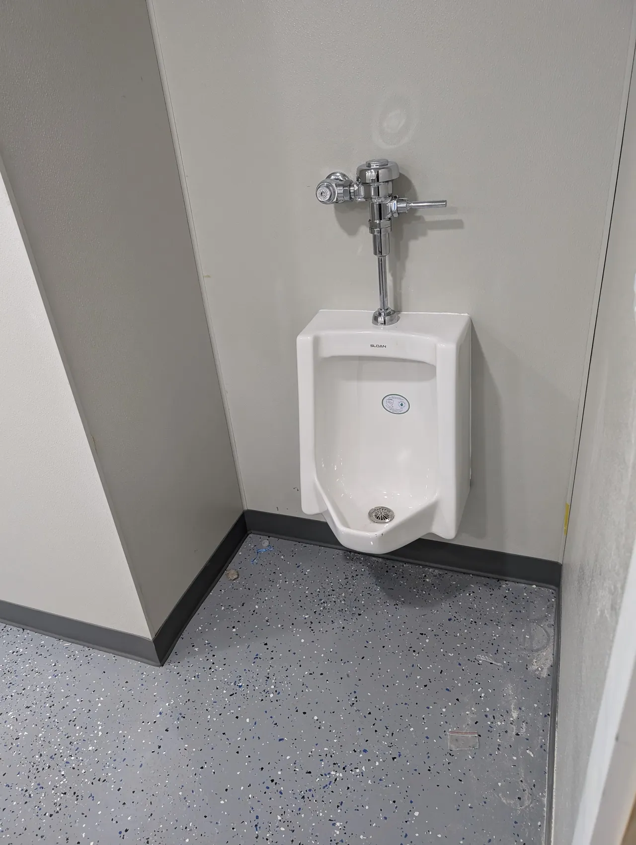 Wall-mounted urinal in a gray restroom corner with speckled floor and black baseboard