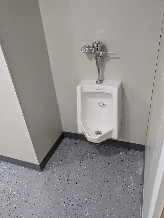 Wall-mounted urinal in a gray restroom corner with speckled floor and black baseboard